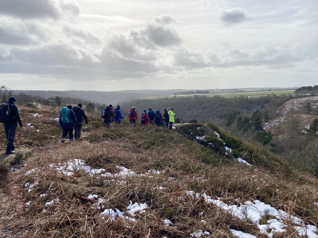 Walkers on bracken covered moorland overlooking a wooded valley, with a dusting of snow on the ground